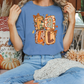 A woman wearing a blue t-shirt with a pumpkin-themed design is sitting on a bench surrounded by fall decorations, including pumpkins and other seasonal items.