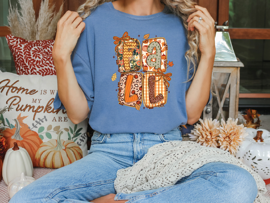 A woman wearing a blue t-shirt with a pumpkin-themed design is sitting on a bench surrounded by fall decorations, including pumpkins and other seasonal items.