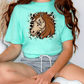 A woman wearing a turquoise t-shirt with a graphic of a horse's head and a patterned design, sitting cross-legged on the floor.