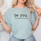 A woman wearing a light blue t-shirt with the text "be you. The world will adjust" printed on it.