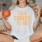 A woman sitting on a bed, holding a mug and wearing a white t-shirt with the text "TALK TURKEY TO ME" printed on it.