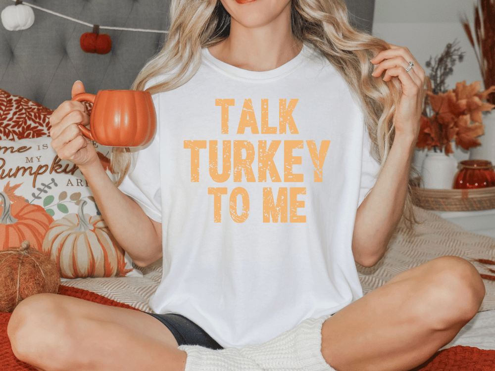 A woman sitting on a bed, holding a mug and wearing a white t-shirt with the text "TALK TURKEY TO ME" printed on it.