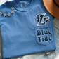 A blue t-shirt with the text "Blue Tide" printed on it, along with a logo, is displayed on a bed with a necklace and a hat.