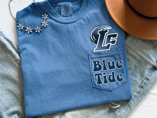 A blue t-shirt with the text "Blue Tide" printed on it, along with a logo, is displayed on a bed with a necklace and a hat.