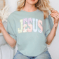 A woman wearing a green t-shirt with the word "JESUS" printed on it, smiling and posing with her hands behind her back.