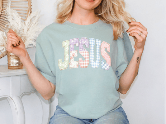A woman wearing a green t-shirt with the word "JESUS" printed on it, smiling and posing with her hands behind her back.