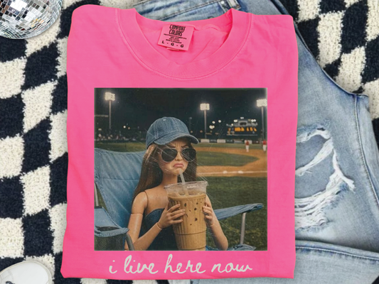 A pink t-shirt with a printed image of a woman wearing sunglasses and holding a drink, sitting on a chair in a field at night.