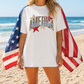 A woman wearing a white t-shirt with an American flag design stands on a beach, with the ocean visible in the background.