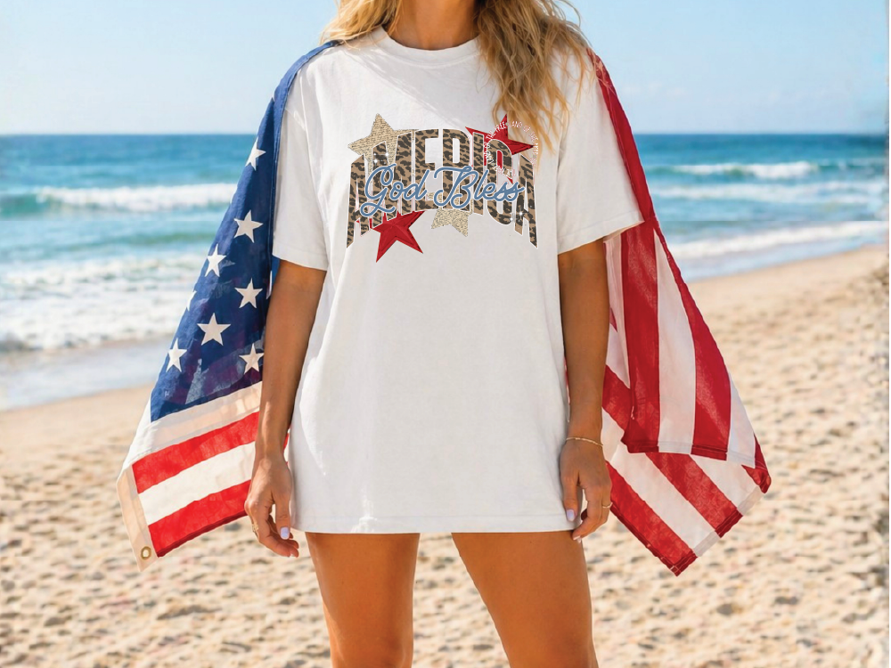 A woman wearing a white t-shirt with an American flag design stands on a beach, with the ocean visible in the background.