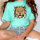 A person wearing a turquoise t-shirt with a leopard print design and gray shorts is sitting cross-legged on the floor.