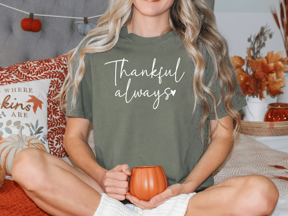 A woman sitting on a bed with a pumpkin-themed pillow and a mug in her hands, wearing a green t-shirt with the text "Thankful always" printed on it.