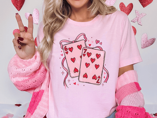 A woman wearing a pink t-shirt with a graphic of playing cards and hearts, making a peace sign with her hand.