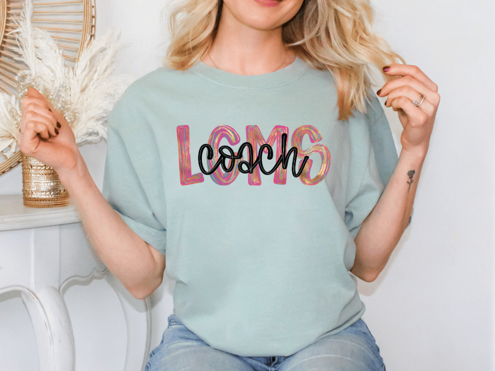 A woman wearing a light blue t-shirt with the word "coach" printed on it, sitting in front of a white wall with a decorative item behind her.