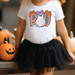 A young girl wearing a white t-shirt with a graphic of a ghost and the word "BOO" is standing next to a table with carved pumpkins.