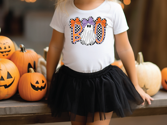 A young girl wearing a white t-shirt with a graphic of a ghost and the word "BOO" is standing next to a table with carved pumpkins.