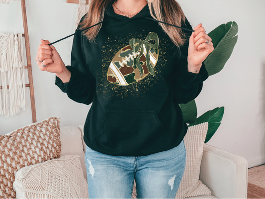 A woman wearing a black hoodie with a football graphic and a green bow, standing in a room with a couch and a plant.