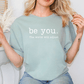 A woman wearing a light blue t-shirt with the text "be you. The world will adjust" printed on it.