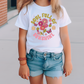 a little girl standing on a sidewalk wearing a shirt with flowers on it
