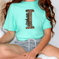 A person wearing a mint green t-shirt with a leopard print design and gray shorts is sitting on the floor.