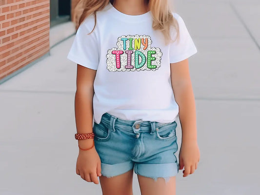 a little girl standing on a sidewalk wearing a t - shirt that says tiny tide