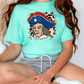 A woman wearing a turquoise t-shirt with a graphic of a man wearing a pirate hat, sitting on the floor.