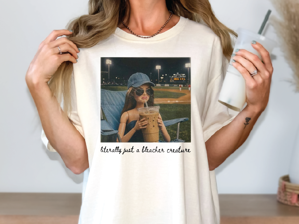 A woman wearing a white t-shirt with a printed image of a woman holding a drink and a caption that reads "Literally just a bleacher creature".