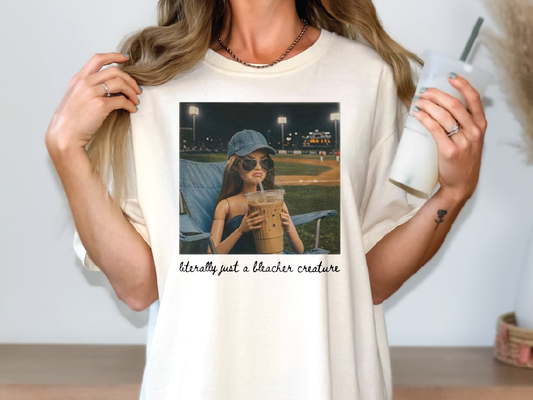 A woman wearing a white t-shirt with a printed image of a woman holding a drink and a caption that reads "Literally just a bleacher creature".