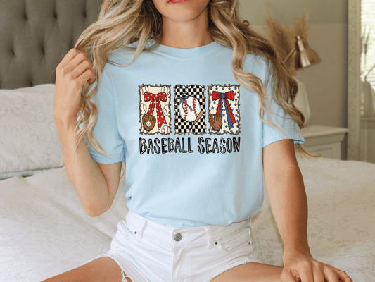 A woman wearing a light blue t-shirt with a baseball-themed design and text, sitting on a bed.