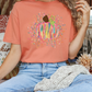 A woman wearing a peach-colored t-shirt with a colorful pumpkin graphic, sitting on a couch with a white blanket and a bouquet of flowers nearby.