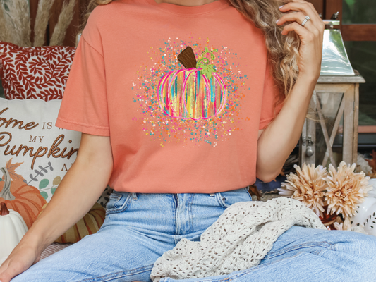A woman wearing a peach-colored t-shirt with a colorful pumpkin graphic, sitting on a couch with a white blanket and a bouquet of flowers nearby.