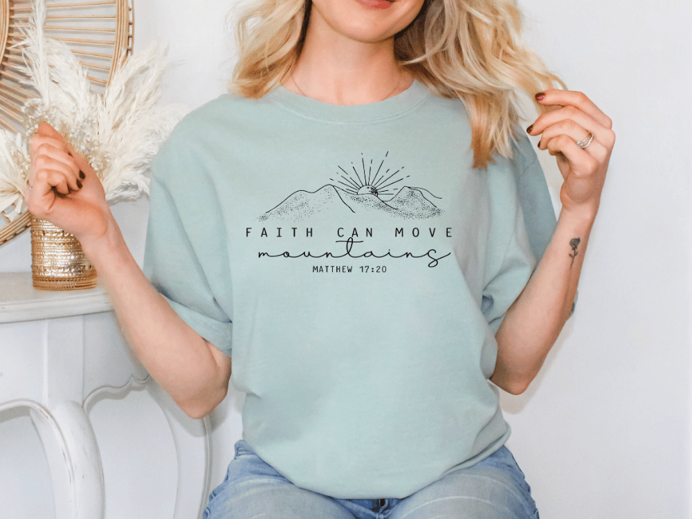 A woman wearing a light blue t-shirt with a graphic design and text, sitting in front of a white wall with a decorative item.