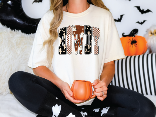A woman wearing a white t-shirt with a Halloween-themed design is sitting on a couch, holding a pumpkin-shaped mug.
