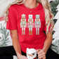 A woman wearing a red t-shirt with a nutcracker design is holding a mug of hot chocolate and surrounded by Christmas decorations, including a Christmas tree and a wreath.