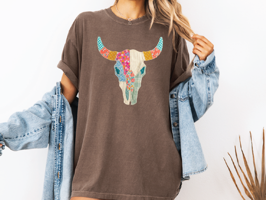 A woman wearing a brown t-shirt with a colorful bull skull design, paired with a denim jacket and denim shorts.