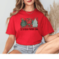 A woman wearing a red t-shirt with a Christmas-themed graphic and text, sitting with her legs crossed.