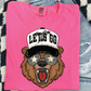 A pink t-shirt with a cartoon bear wearing sunglasses and a hat with a star design, along with the text "LET'S GO" printed on it.