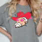A woman wearing a gray t-shirt with a graphic design of a man wearing a red hat and sunglasses, surrounded by pink stars.
