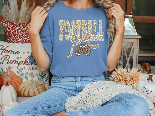 A woman wearing a blue t-shirt with a football graphic and a ribbon, sitting on a couch surrounded by fall decorations.