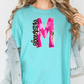 A woman wearing a turquoise t-shirt with a large pink and purple letter "M" printed on it, along with a denim jacket.