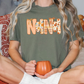 A person sitting on a bed, wearing a green t-shirt with the word "Nana" printed on it, holding a pumpkin-shaped mug.