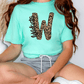 A person wearing a turquoise t-shirt with a leopard print design and gray shorts is sitting on the floor.