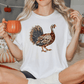 A woman wearing a white t-shirt with a turkey design is sitting on a bed, holding a mug and surrounded by fall decorations.