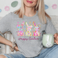 A woman wearing a gray t-shirt with a colorful Easter bunny design and holding a mug, sitting on a couch with a potted plant in the background.