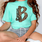 A person wearing a turquoise t-shirt with a leopard print design and gray shorts is sitting on the floor.