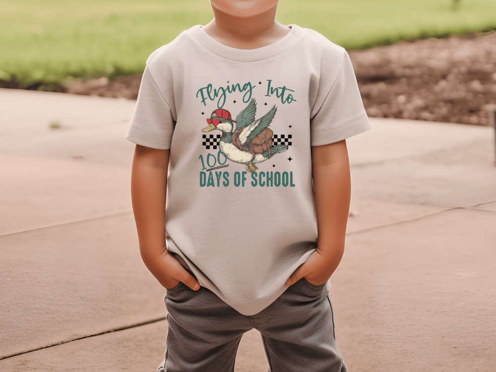 A child wearing a white t-shirt with a graphic design of a duck and the text "Flying into 100 days of school" printed on it.