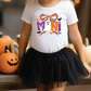 A young girl wearing a white t-shirt with a Halloween-themed design and a black tulle skirt stands in front of a collection of carved pumpkins.
