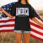 A person standing in a field holding an American flag, wearing a black t-shirt with the text "AMERICA 250 years of freedom" printed on it.