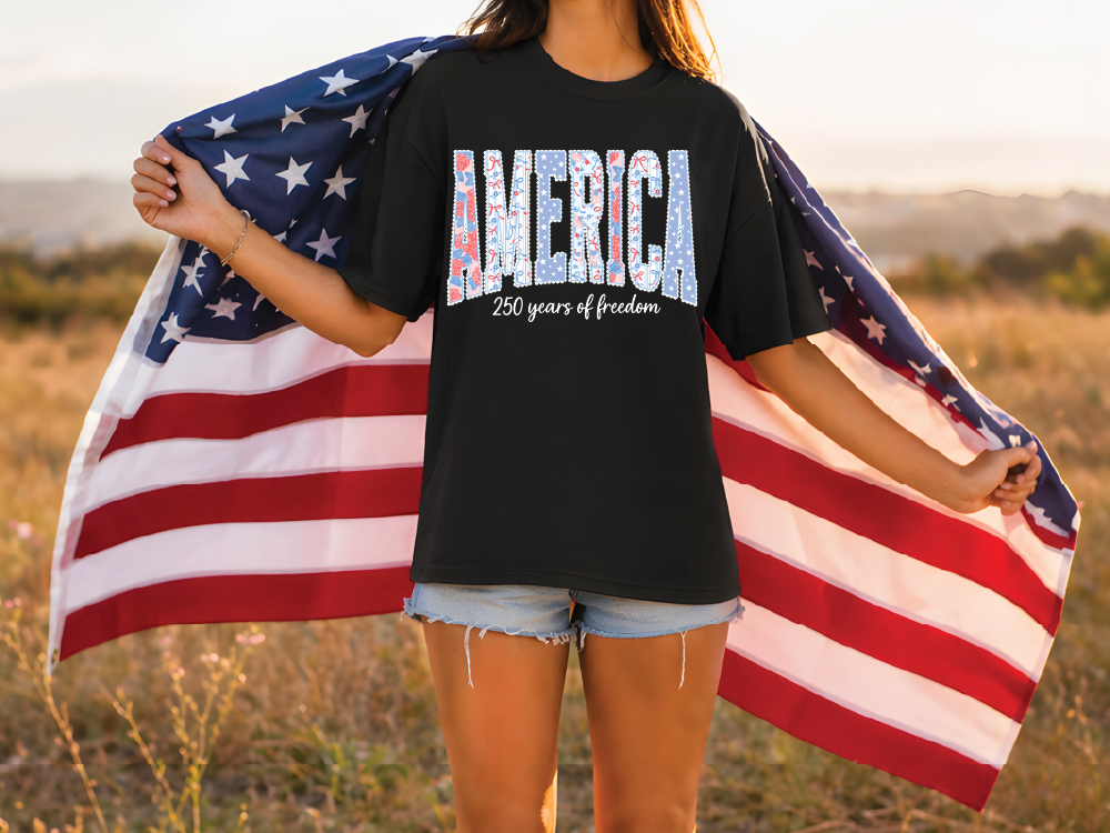 A person standing in a field holding an American flag, wearing a black t-shirt with the text "AMERICA 250 years of freedom" printed on it.