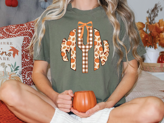 A woman is sitting on a couch, wearing a green t-shirt with a graphic design of a pumpkin and autumn leaves. She is holding a mug in her hands.