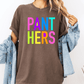 A person wearing a brown t-shirt with the text "PANT HERS" in colorful letters, along with a denim jacket, stands against a white background.